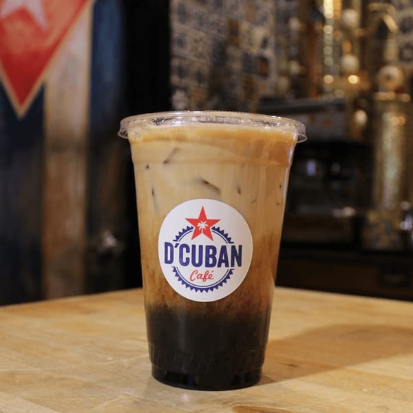 Iced coffee with milk and a D'Cuban Cafe logo sticker on a wooden table.