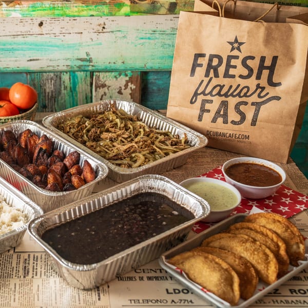 Cuban takeout meal with plantains, rice, black beans, empanadas, and sauces in foil containers.