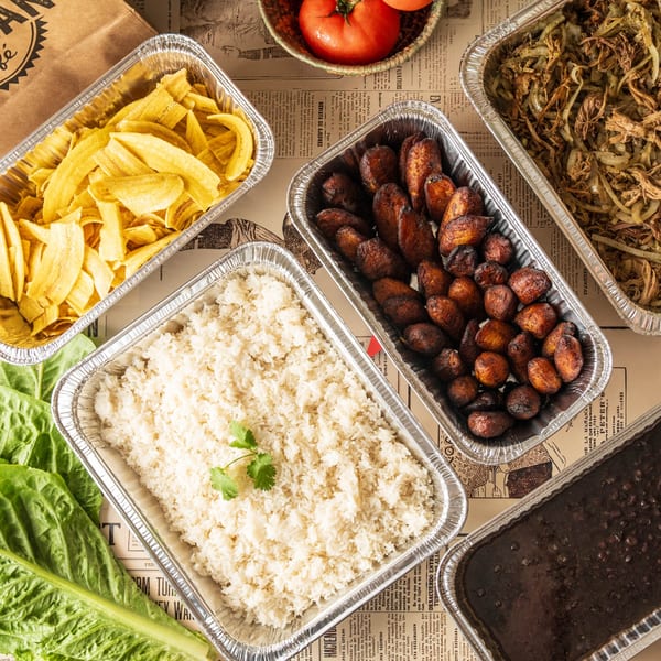 Cuban dishes in foil containers: plantain chips, white rice, black beans, fried plantains, and shredded beef.