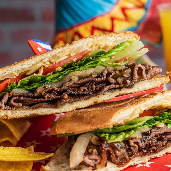 Two halves of a D'Cuban Cafe steak sandwich with lettuce, tomato, and onions, with plantain chips.