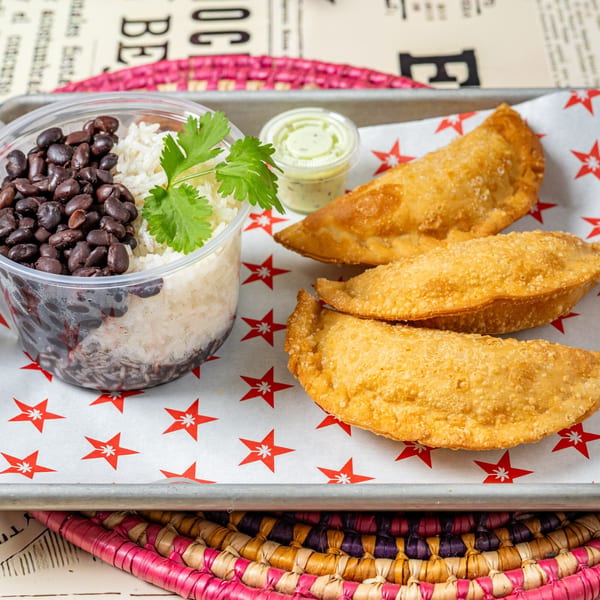 Cuban black beans and rice in a clear container with three golden empanadas and a dipping sauce.