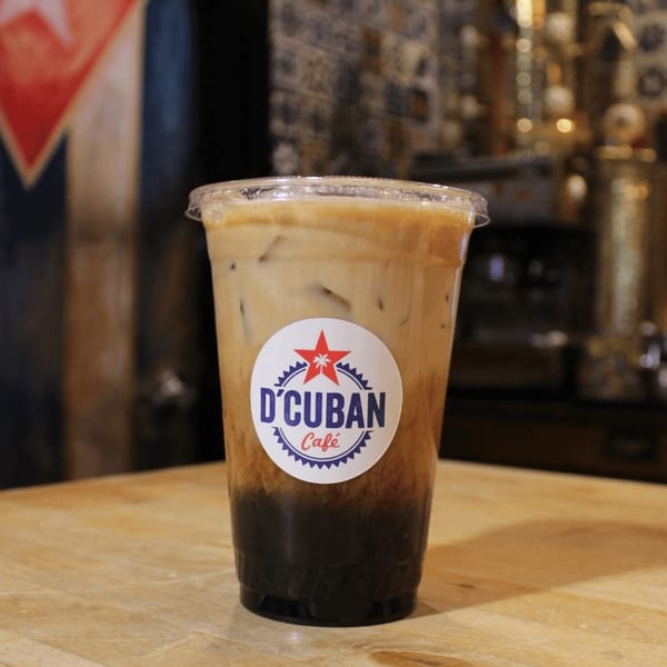 Iced coffee with milk and D'Cuban Cafe logo on a wooden table with a Cuban flag in the background.