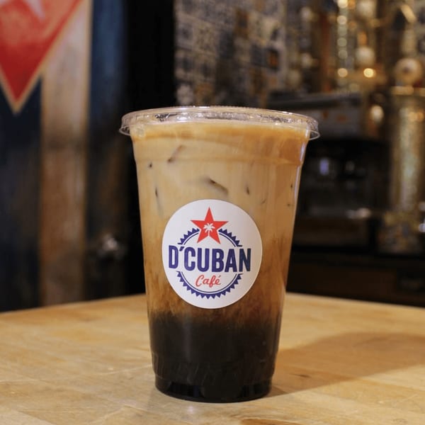 Iced coffee with milk and a D'Cuban Cafe logo sticker on a wooden table.