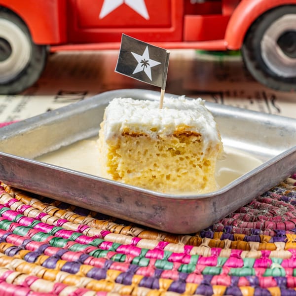 A slice of tres leches cake with a Cuban flag toothpick, in a metal tray.