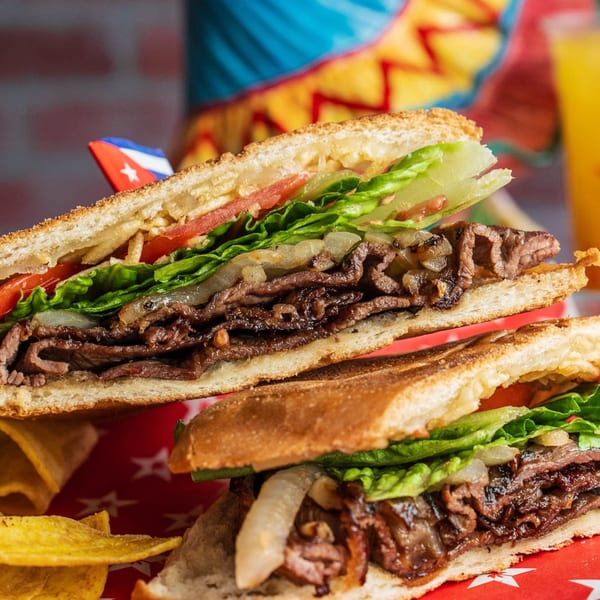 Two halves of a Cuban steak sandwich with lettuce, tomato, and onions, with plantain chips.