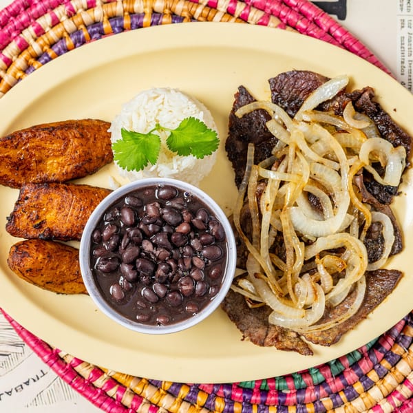 Cuban steak with sautéed onions, white rice, black beans, and fried plantains on a yellow plate.