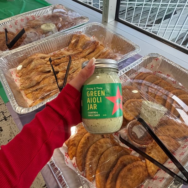 A hand holds a jar of D'Cuban Cafe's green aioli over trays of empanadas and pastries.