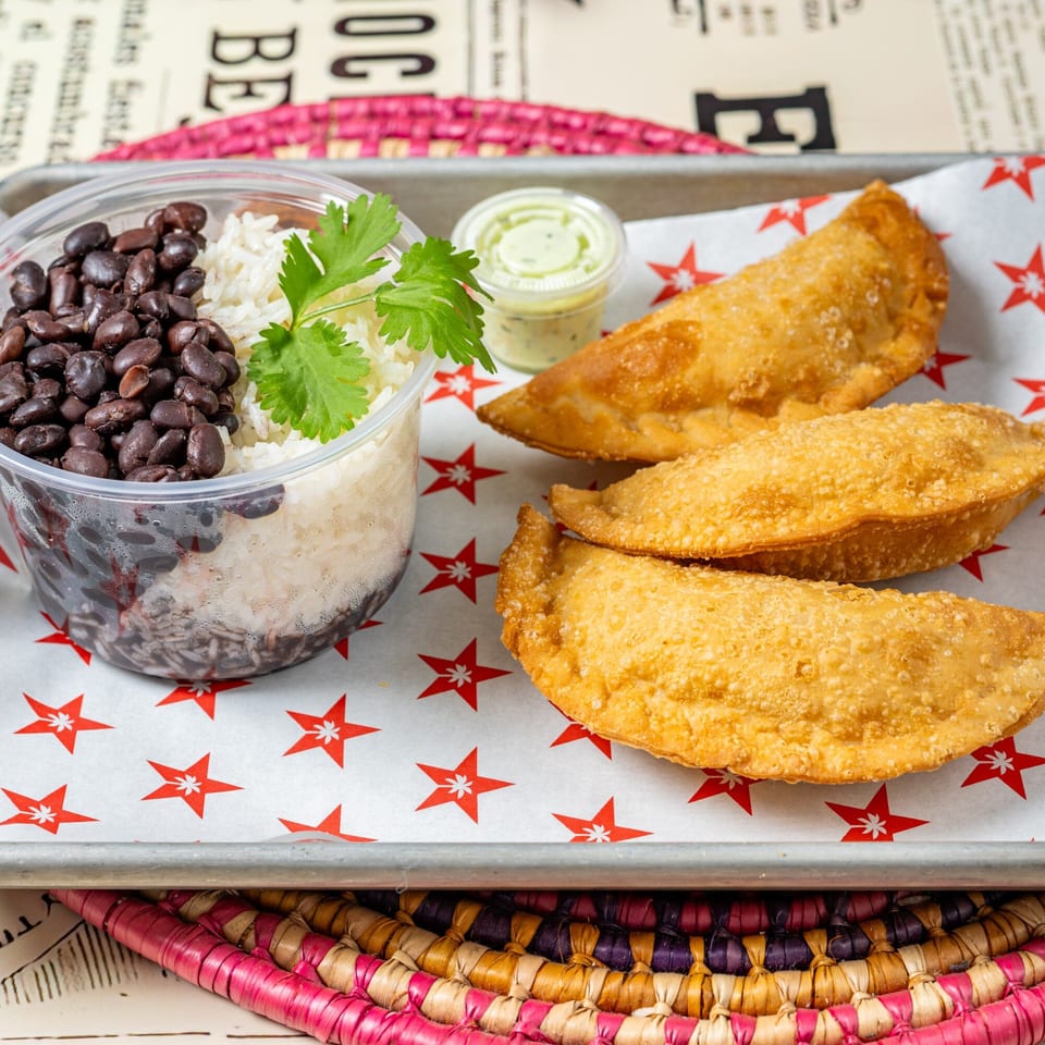 Cuban meal with black beans, white rice, cilantro, three empanadas, and a side of green sauce.